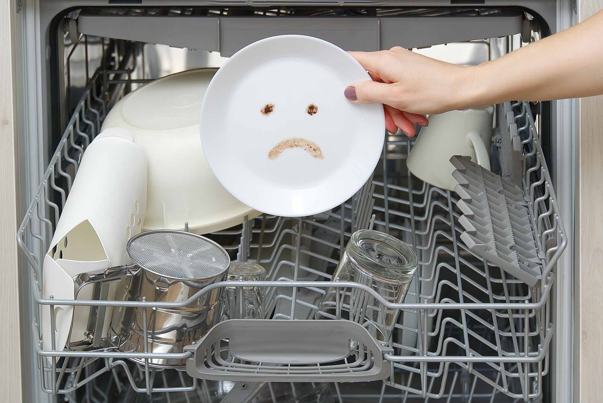 A dishwasher full of half-cleaned dishes. A hand holds up a plate with a frowning face drawn on it in dirt